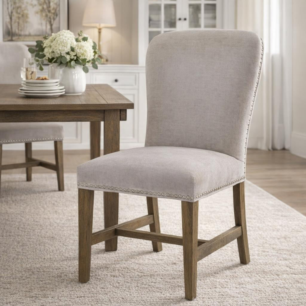 Pale grey stud-edged dining chair on a light rug beside a brown dining table set with stacked white plates and a vase of white flowers.