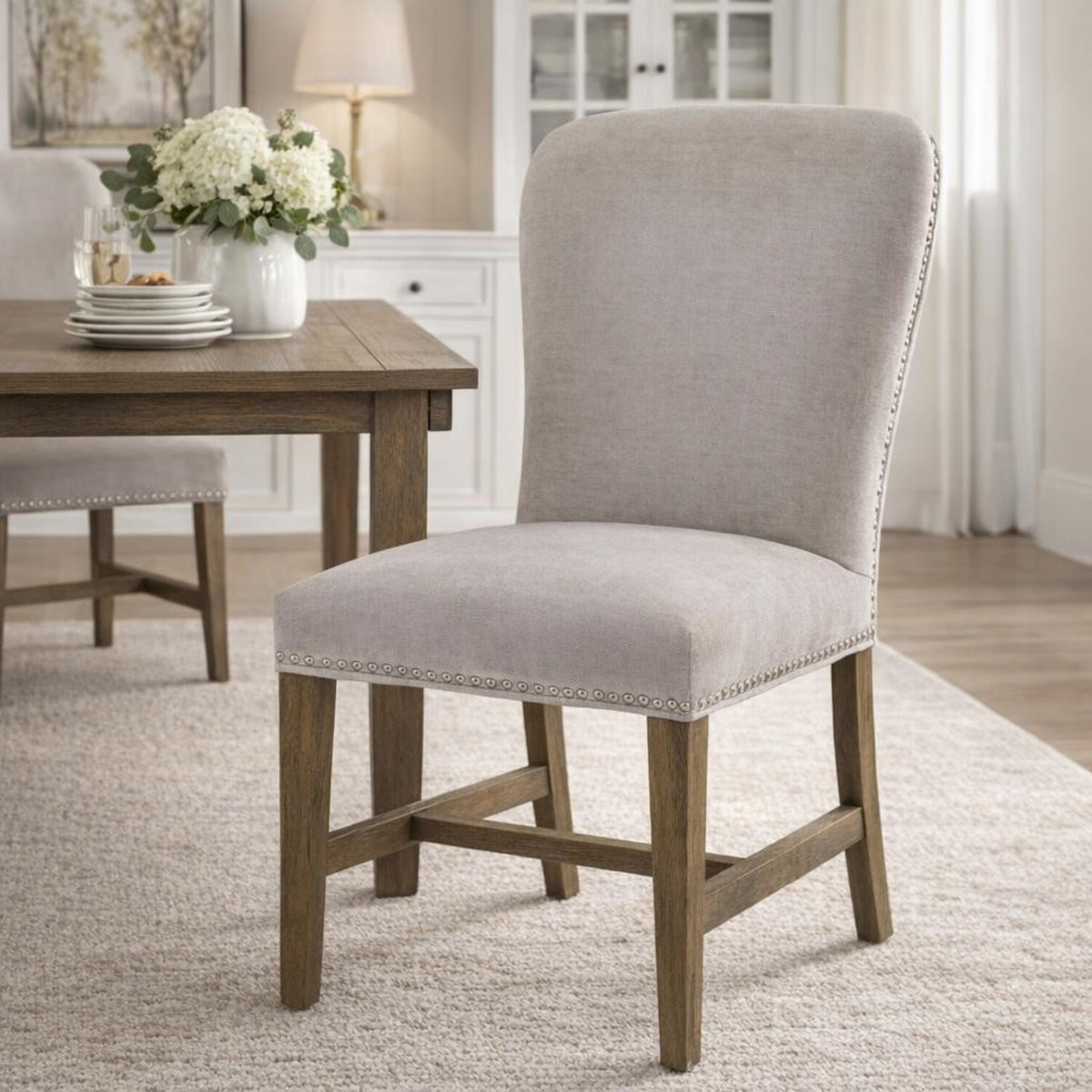 Pale grey stud-edged dining chair on a light rug beside a brown dining table set with stacked white plates and a vase of white flowers.