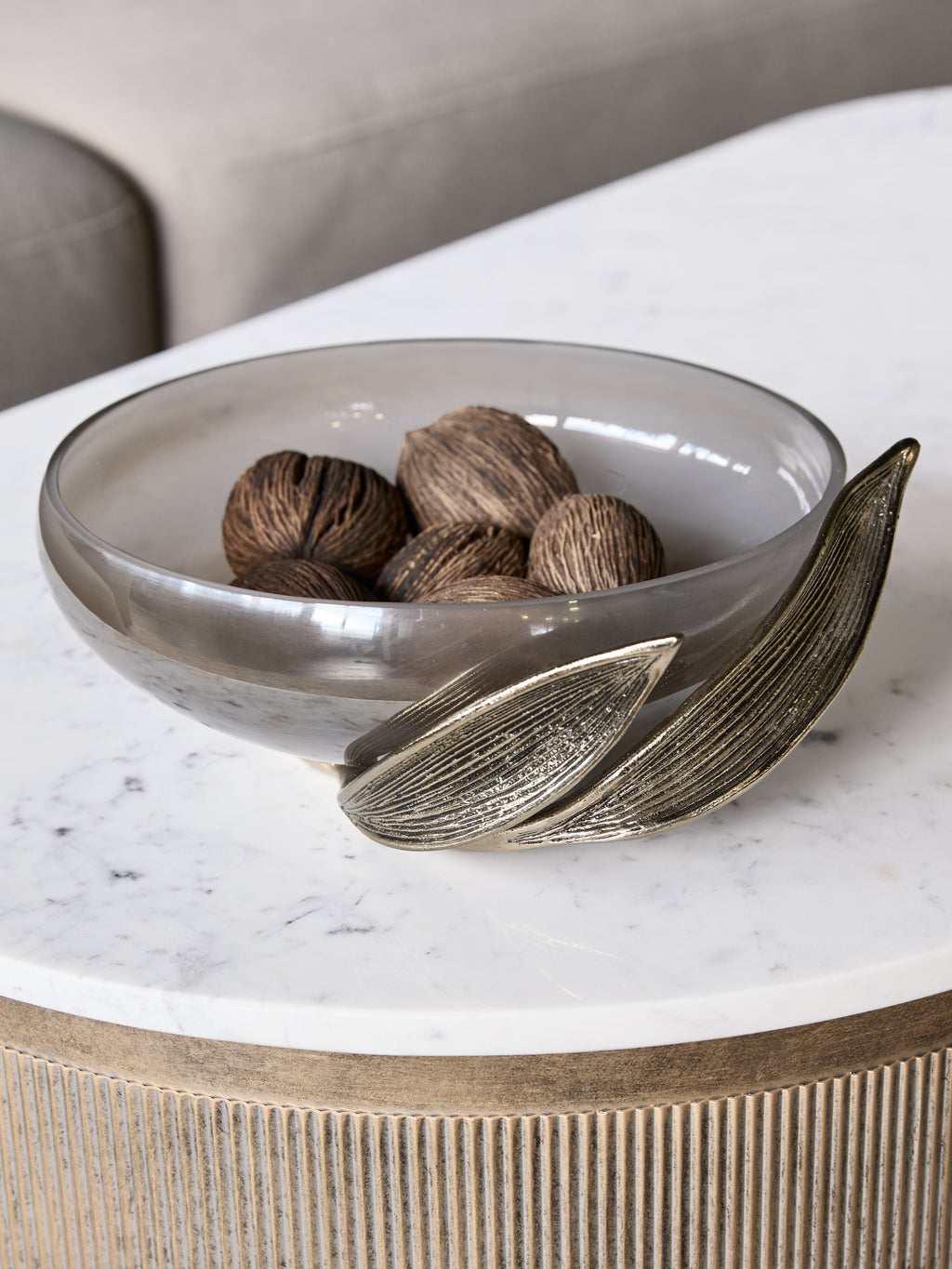 Leaf-framed glass bowl filled with brown decorative balls on a white veined coffee table beside a neutral sofa.