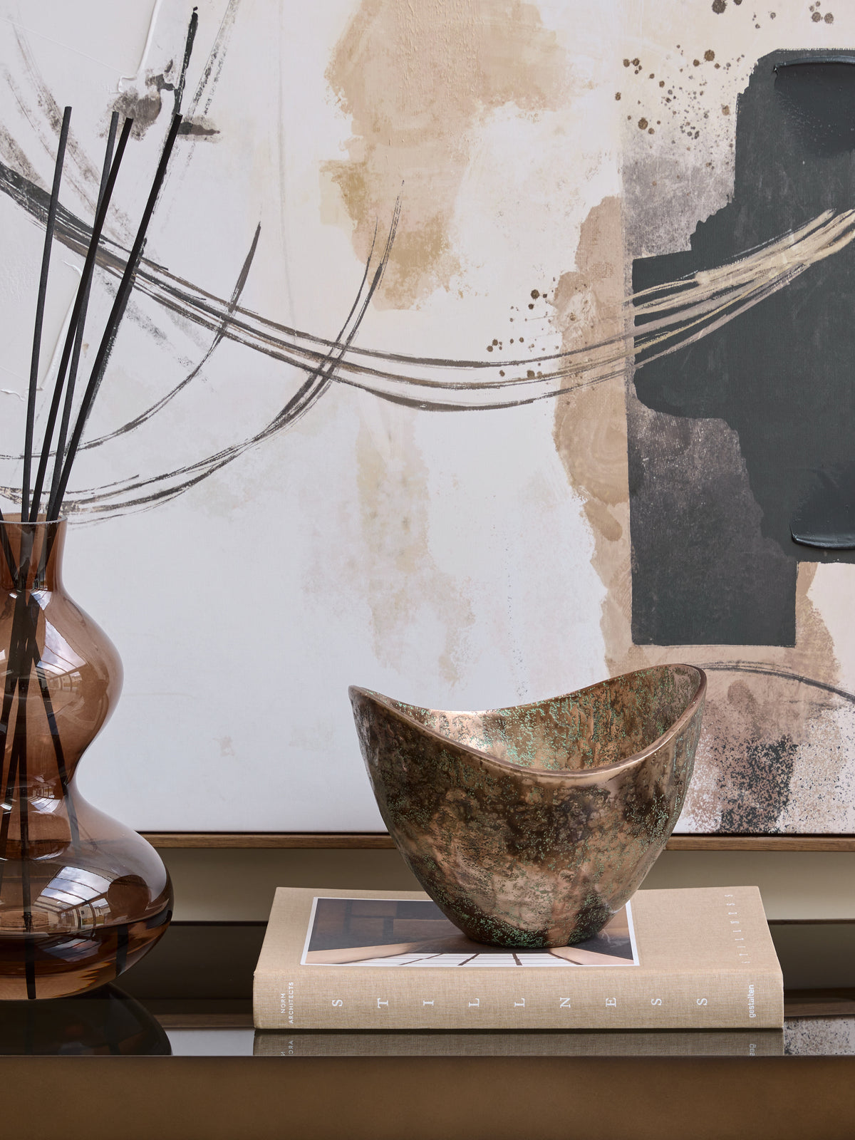 Sweeping-rim bowl displayed on a beige book beside an amber glass vase, with abstract artwork behind.