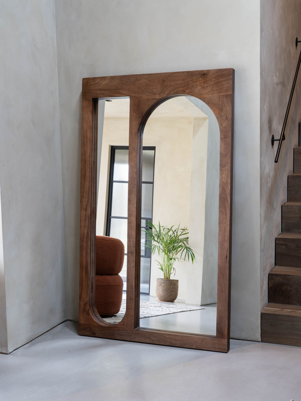 Two-pane wooden floor mirror leaning against a pale hallway wall beside stairs, reflecting a plant and rust-coloured chair.