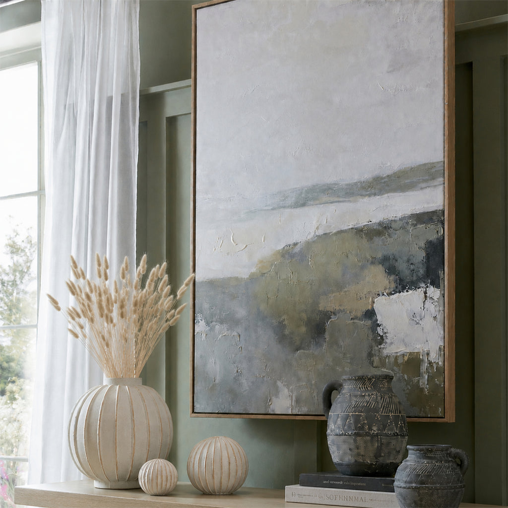Pale botanical plumes displayed in a ribbed cream vase beside matching round ornaments, dark jugs and framed artwork against a green wall.