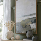 Pale botanical plumes displayed in a ribbed cream vase beside matching round ornaments, dark jugs and framed artwork against a green wall.