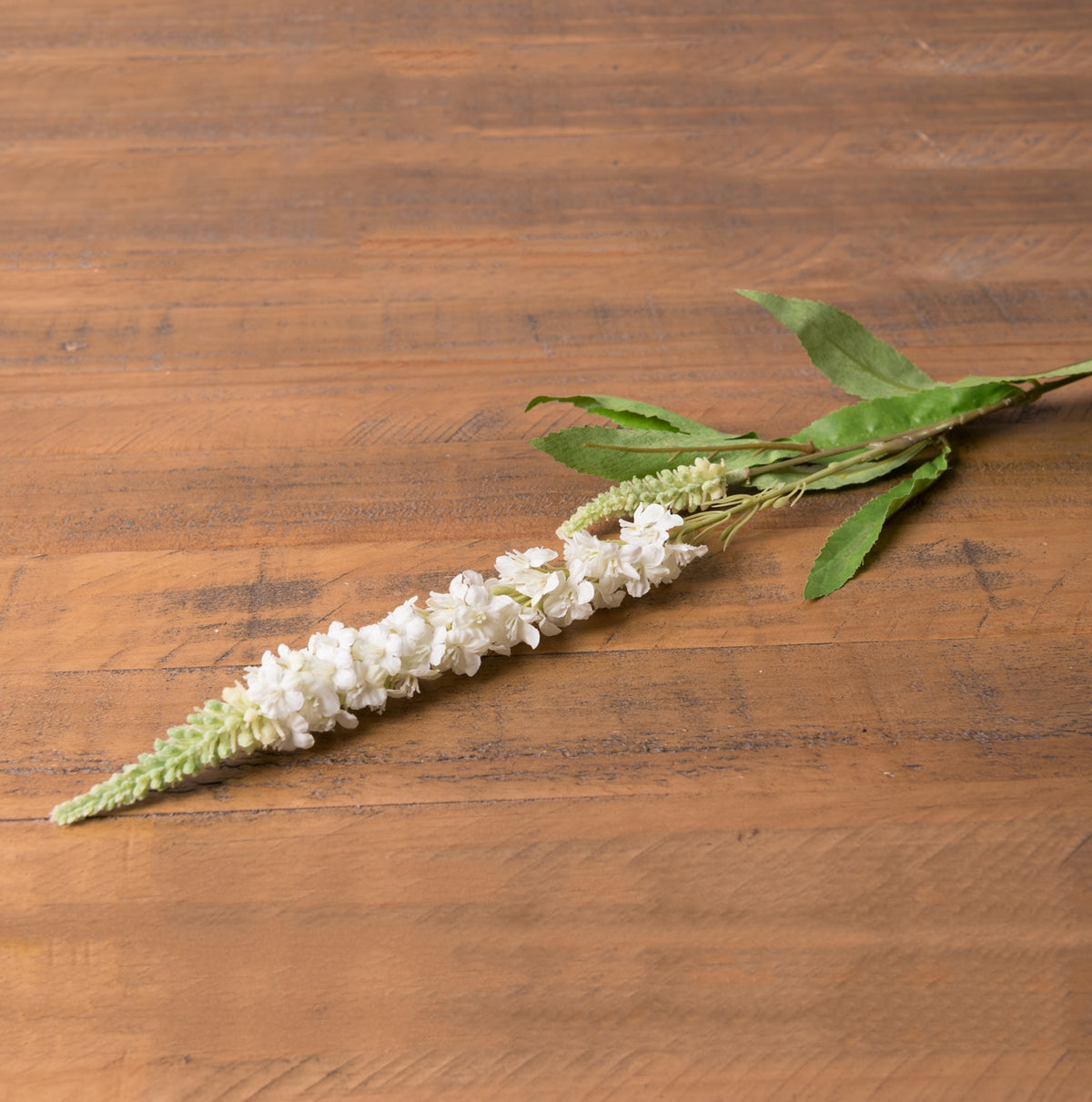 Artificial flower stem lying diagonally on a wooden surface, showing small white florets along the spike, pale-green buds at the tip and green leaves behind.