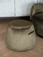 Green tapered stool on a wood floor beside a similarly coloured upholstered armchair, with a patterned cabinet behind.