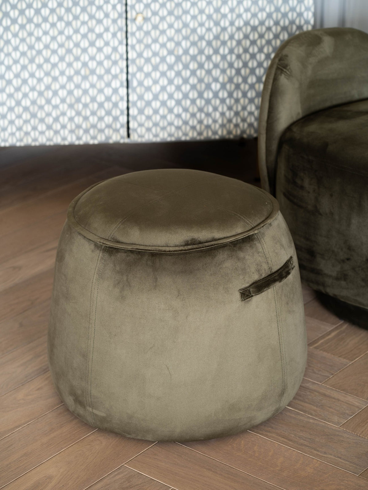 Green tapered stool on a wood floor beside a similarly coloured upholstered armchair, with a patterned cabinet behind.