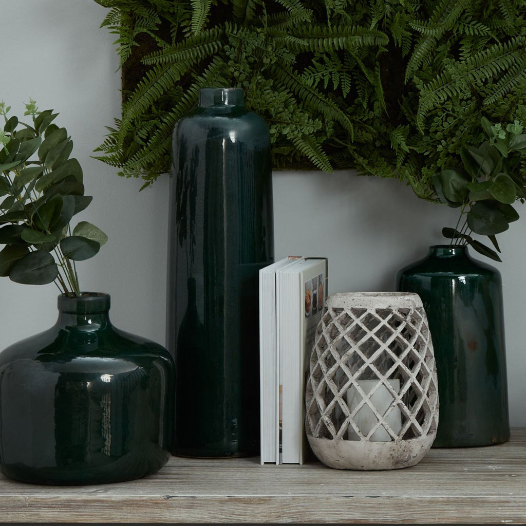 Pale lattice lantern with an unlit white candle visible inside, styled on a wooden surface beside a book and dark green vases beneath fern foliage.
