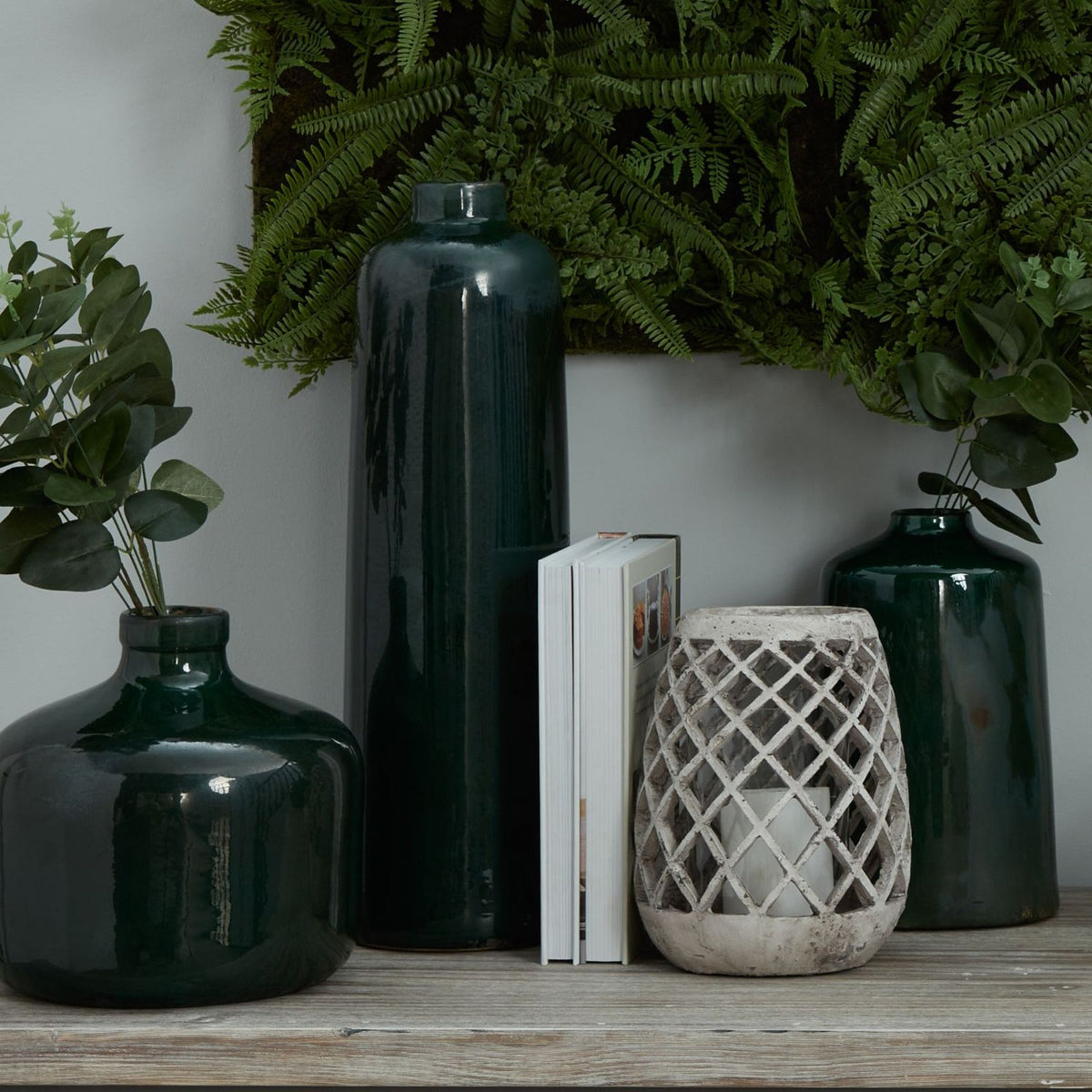 Pale lattice lantern with an unlit white candle visible inside, styled on a wooden surface beside a book and dark green vases beneath fern foliage.