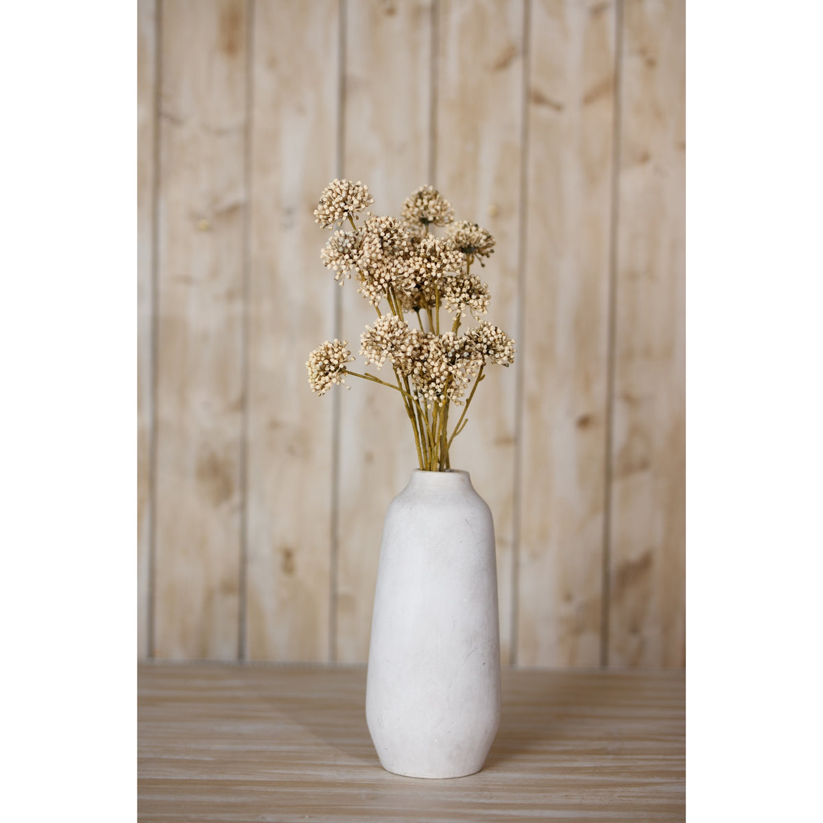 Botanical sprays arranged in a tall off-white vase on a wooden surface in front of pale vertical wood panelling.