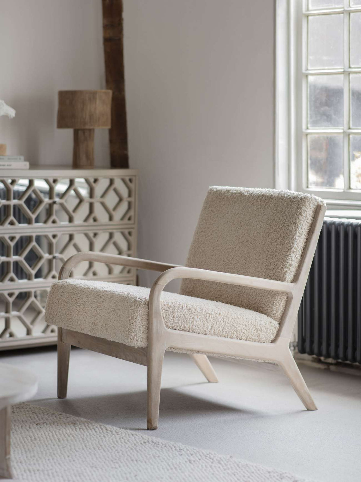 Teddy chair beside a window in a neutral room, with a mirrored cabinet and woven rug nearby.