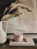 Pink marble storage jar on a book beside a pale textured vase with dried leaves, in front of a neutral abstract artwork.