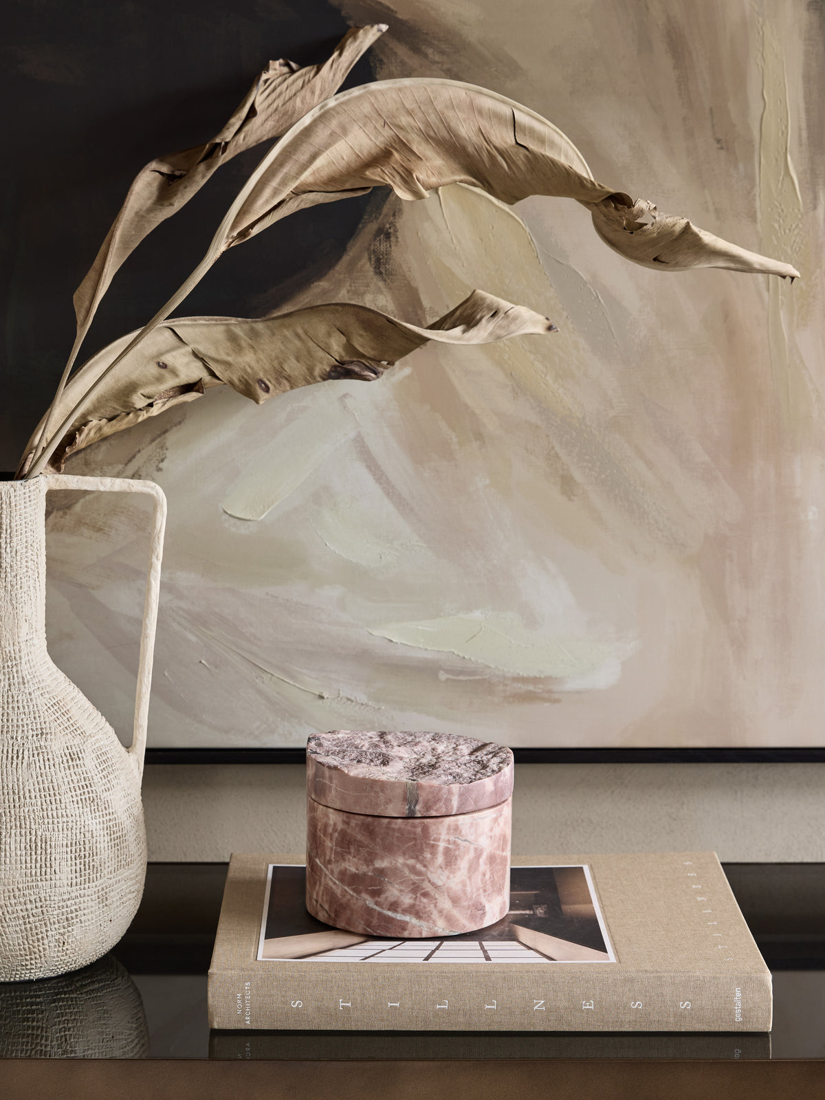 Pink marble storage jar on a book beside a pale textured vase with dried leaves, in front of a neutral abstract artwork.