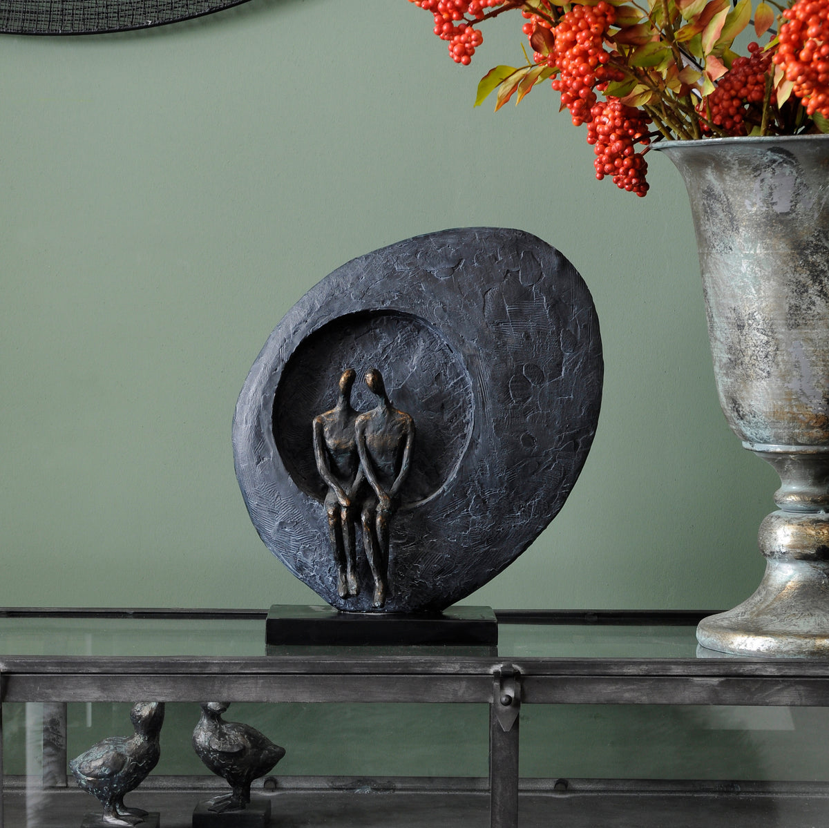 Accord Sculpture displayed on a glass-topped metal cabinet against a green wall, beside a tall urn holding red berries.