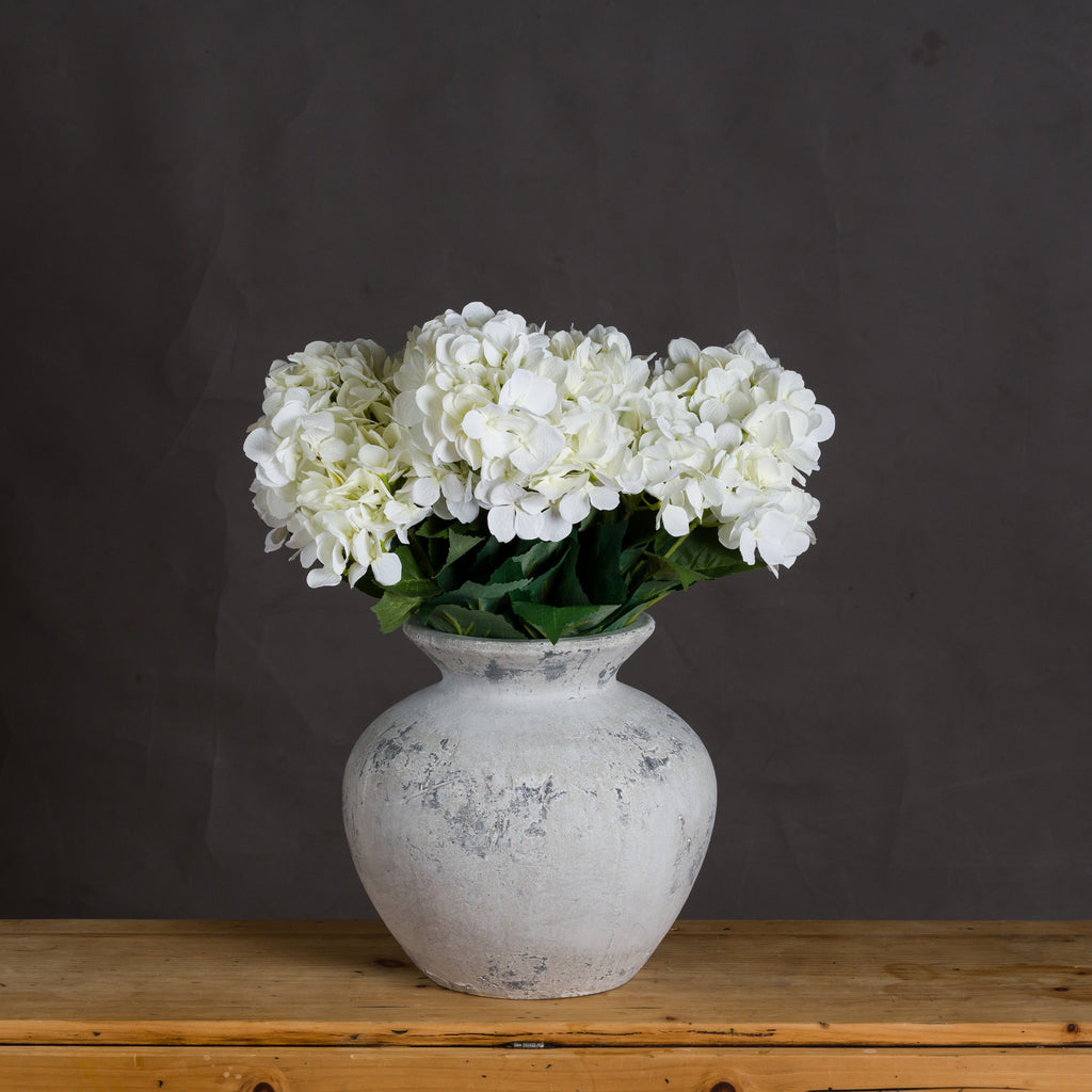 Thicklip Vase styled with white hydrangea blooms and green leaves on a wooden surface against a dark grey wall.