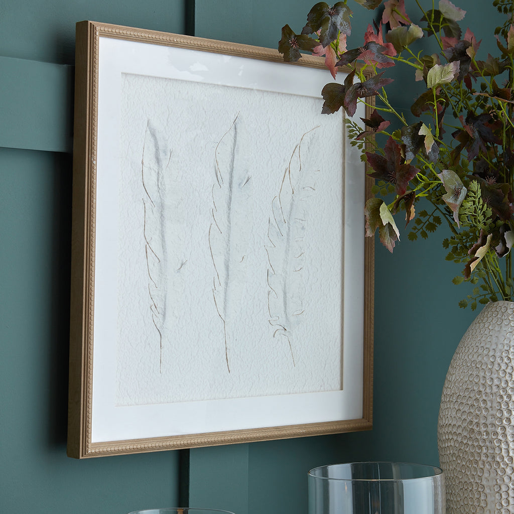 Three Feathers Framed Artwork displayed against a teal panelled wall beside leafy stems in a textured pale vase.
