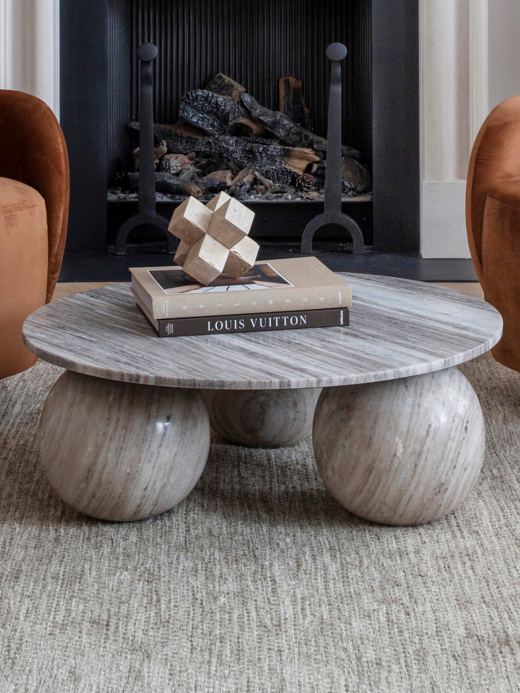Marble orb coffee table styled with books and a geometric ornament between rust-coloured chairs in front of a fireplace.