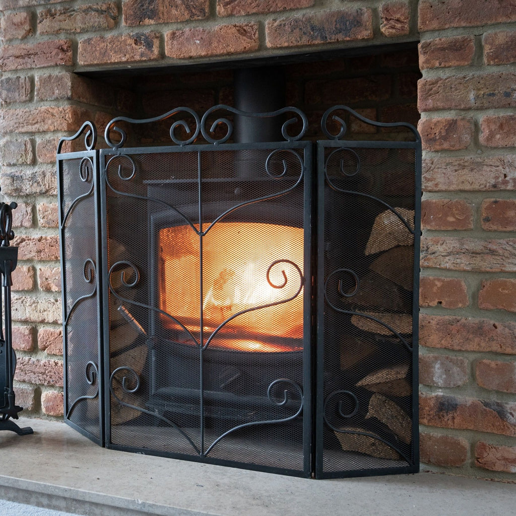 Black three-panel scrollwork fire screen on a pale hearth in front of a glowing stove within a brick fireplace, with fireplace tools partly visible to the left.