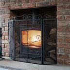 Black three-panel scrollwork fire screen on a pale hearth in front of a glowing stove within a brick fireplace, with fireplace tools partly visible to the left.