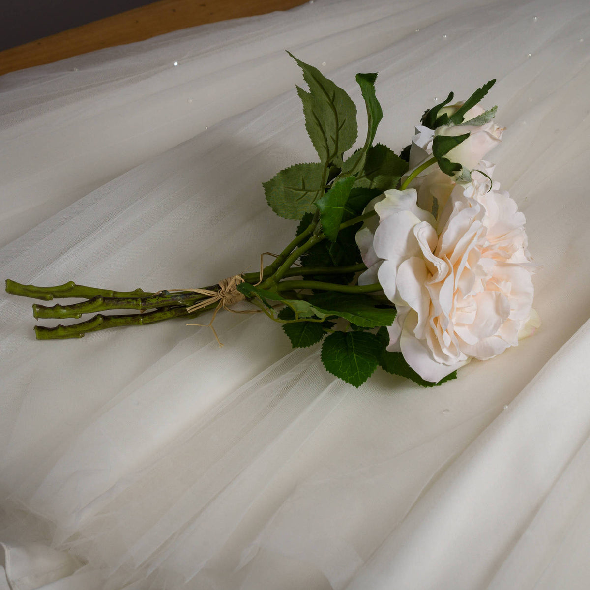 Tied artificial rose bouquet lying diagonally on sheer white fabric, with bundled stems to the left and pale flowers and green foliage to the right.