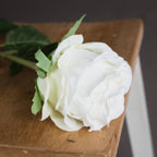 Close-up of the artificial white rose resting on a wooden surface, showing overlapping petals and pointed green sepals around the flower head.