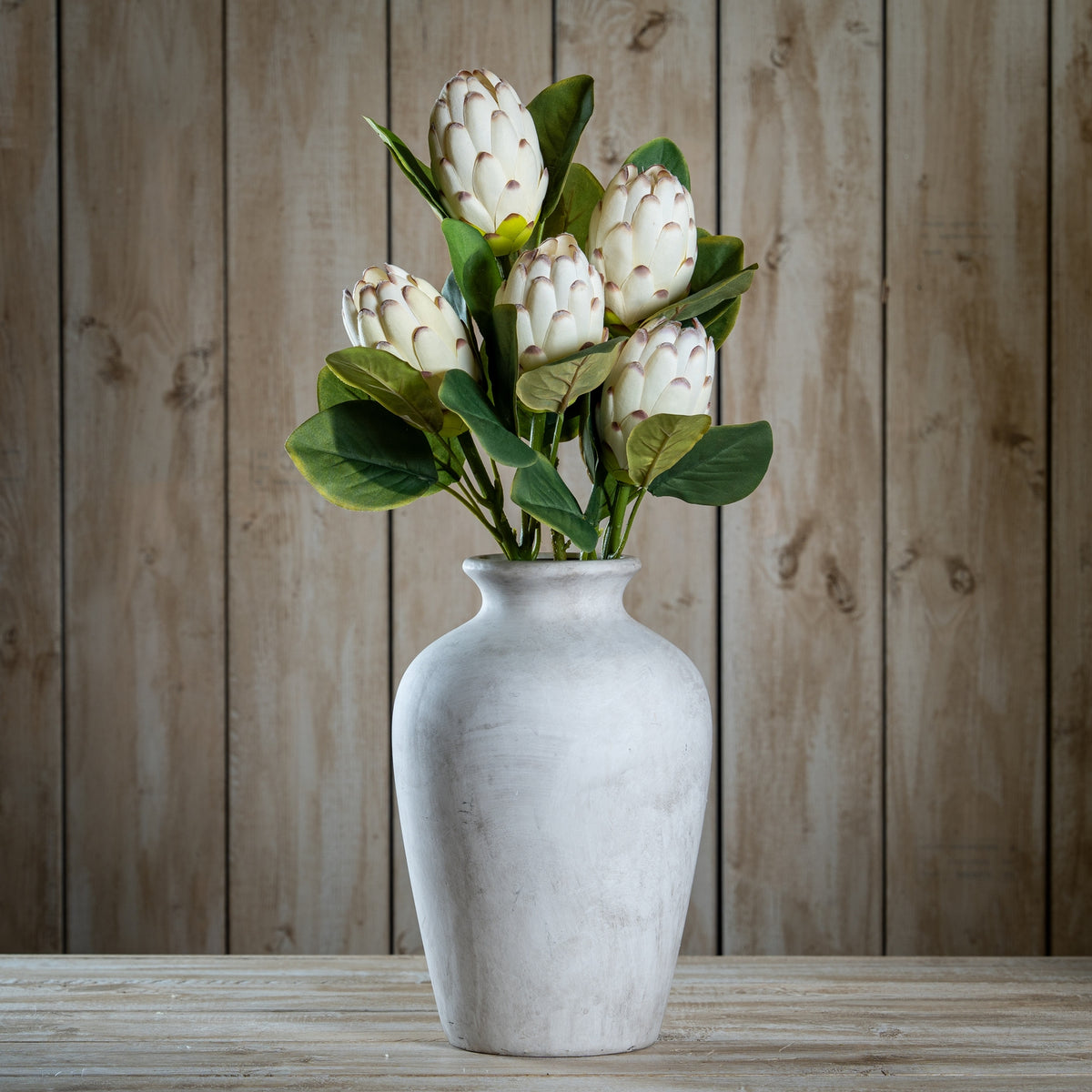 Cream-white tipped cone artificial flowers and broad green leaves arranged in a pale rounded vase on a wooden surface against vertical wood panelling.
