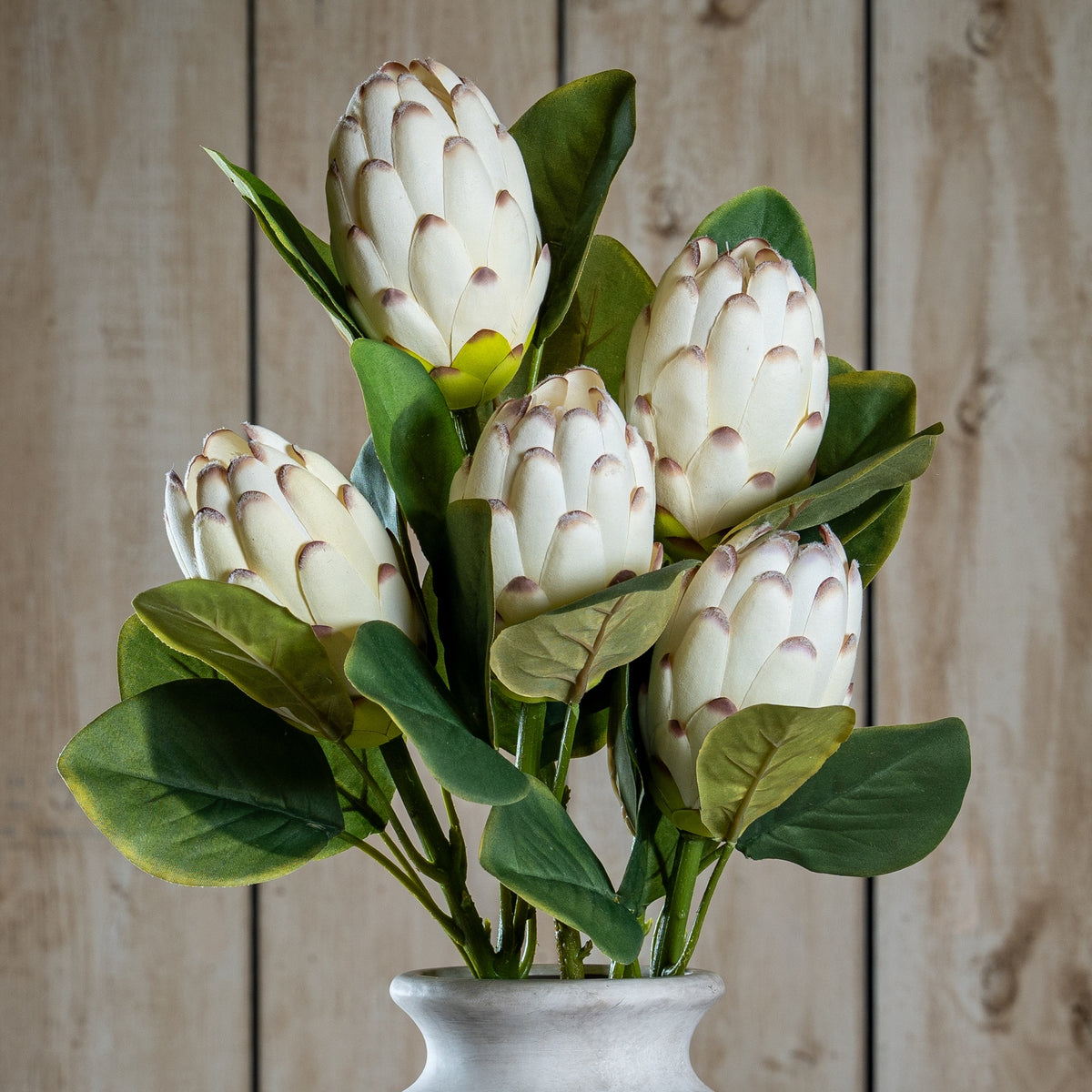 Close-up of cream-white cone-shaped artificial flower heads with overlapping mauve-brown-tipped bracts, veined green leaves and stems above a pale vase rim.