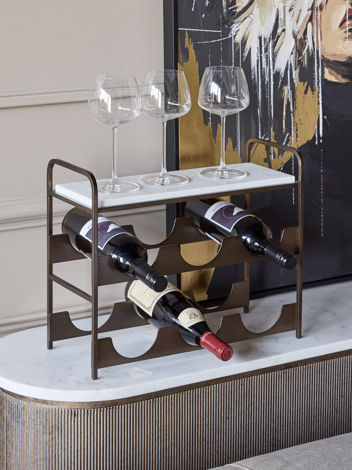 Wine rack holding three bottles with three wine glasses on its marble top, styled on a sideboard in front of abstract artwork.
