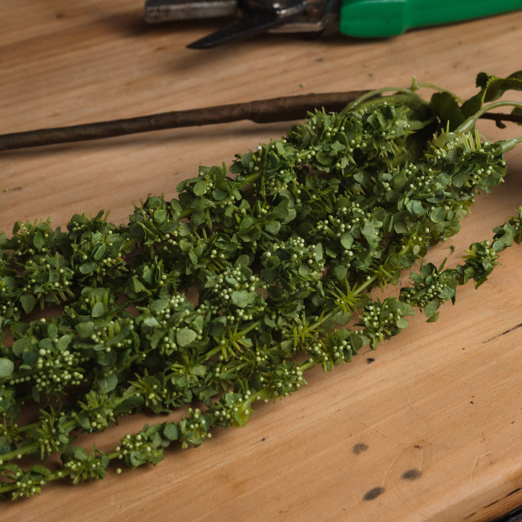 Close-up of the artificial amaranthus strands on a wooden surface, showing densely clustered green details with small rounded buds.