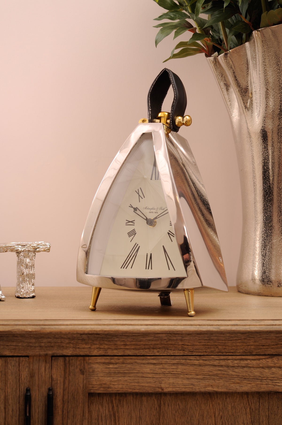Triangular silver clock on a wooden sideboard beside a tall metallic vase of foliage, showing its wedge-shaped side and brass feet.