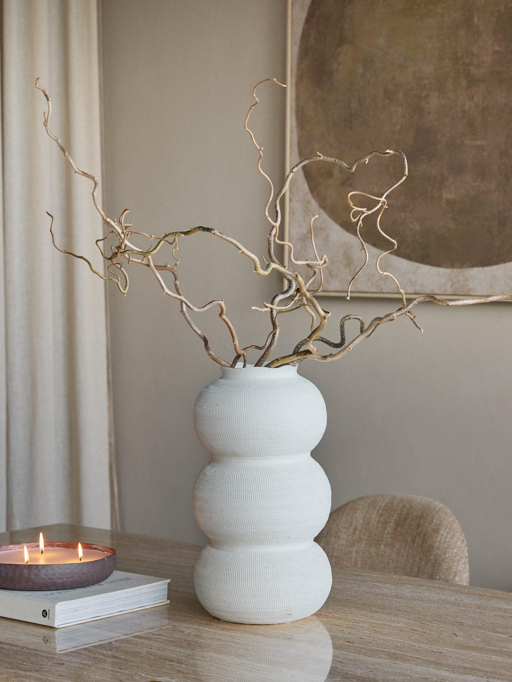 White triple curve vase holding tall twisting branches on a table beside a lit candle and book, with a chair and artwork behind.