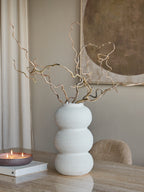 White triple curve vase holding tall twisting branches on a table beside a lit candle and book, with a chair and artwork behind.
