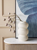 White triple curve vase holding arching decorative stems on a pale console top, in front of blue abstract artwork and a cream panelled wall.