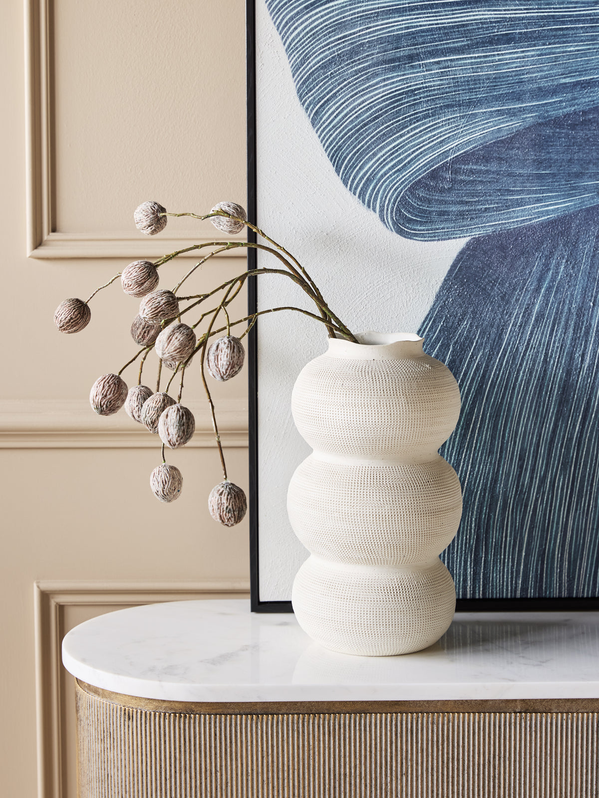 White triple curve vase holding arching decorative stems on a pale console top, in front of blue abstract artwork and a cream panelled wall.