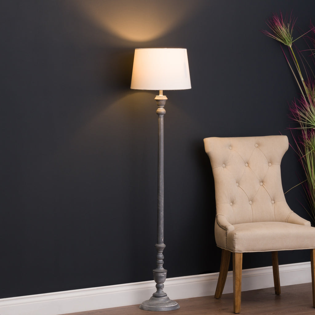 Turned Interval Floor Lamp pictured lit beside a pale buttoned chair against a dark wall, with tall decorative stems to the right.