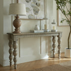 Console table against a cream panelled wall, styled with a lamp, books, a pear ornament and two glass lanterns beneath botanical artwork, beside a potted tree.