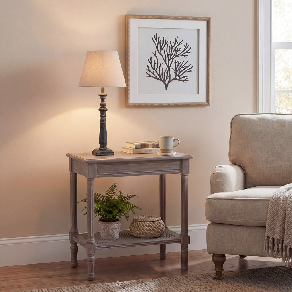 Table lamp pictured with its shade glowing on a side table beside books and a mug, beneath a framed branching print and next to a pale armchair.