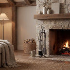 Twist & Knot fireplace tool set beside a lit stone fireplace, with a woven log basket, timber mantel and neutral furnishings.