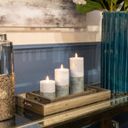 Styled scene with three heights of two-tone grey candles and bright flame-shaped elements on wooden trays, beside a tall teal ribbed vase and metallic vessels on a reflective tabletop.