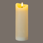 Cream cylindrical LED candle photographed illuminated against a grey background, showing a yellow glow around its recessed top and bright flame-shaped centre.