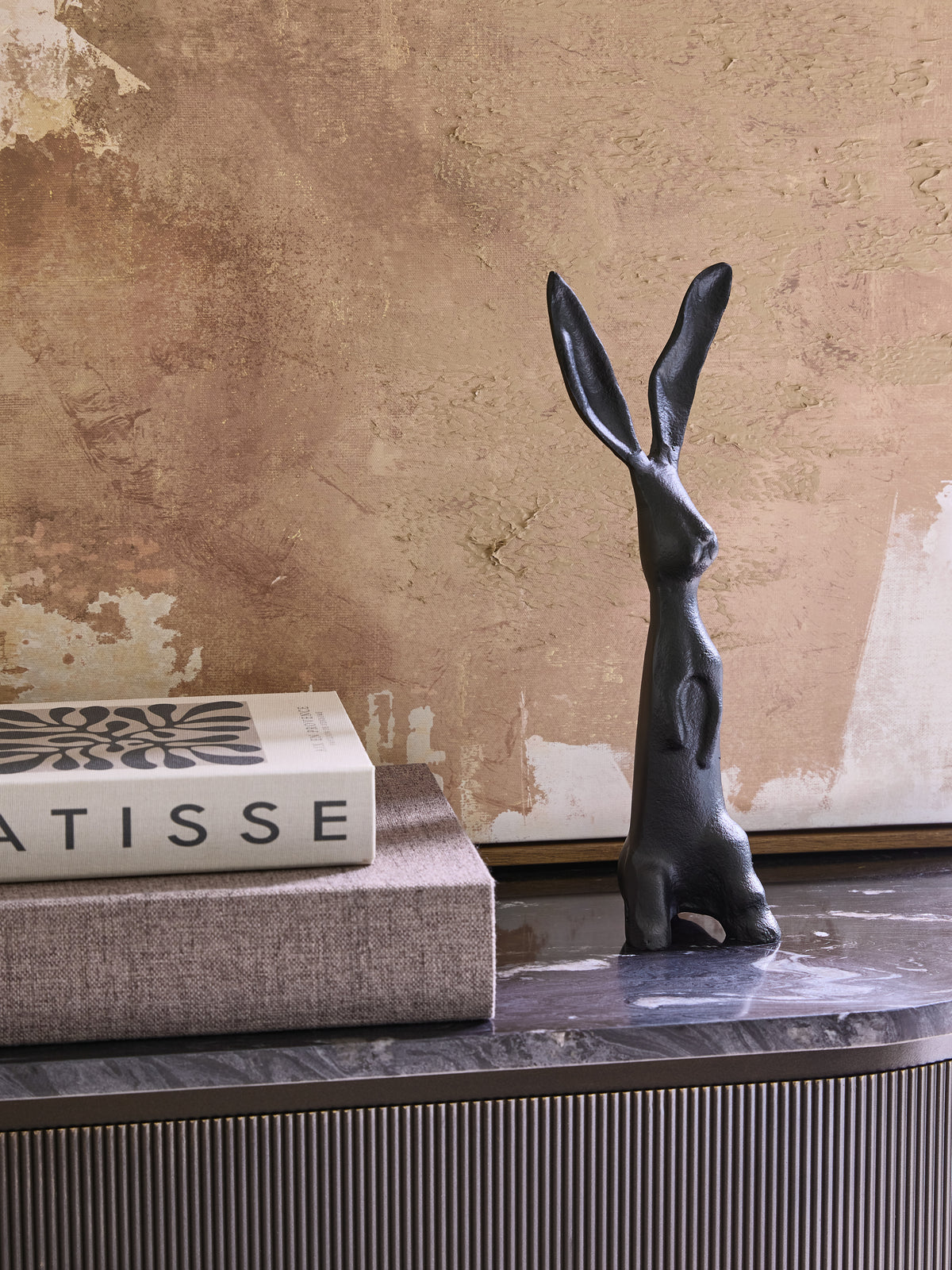Black upright hare sculpture beside stacked books on a dark veined console, with textured beige artwork behind.