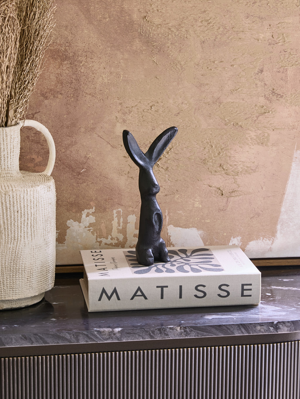 Black hare sculpture displayed on a Matisse book beside a cream jug with dried stems, against a textured warm-toned backdrop.