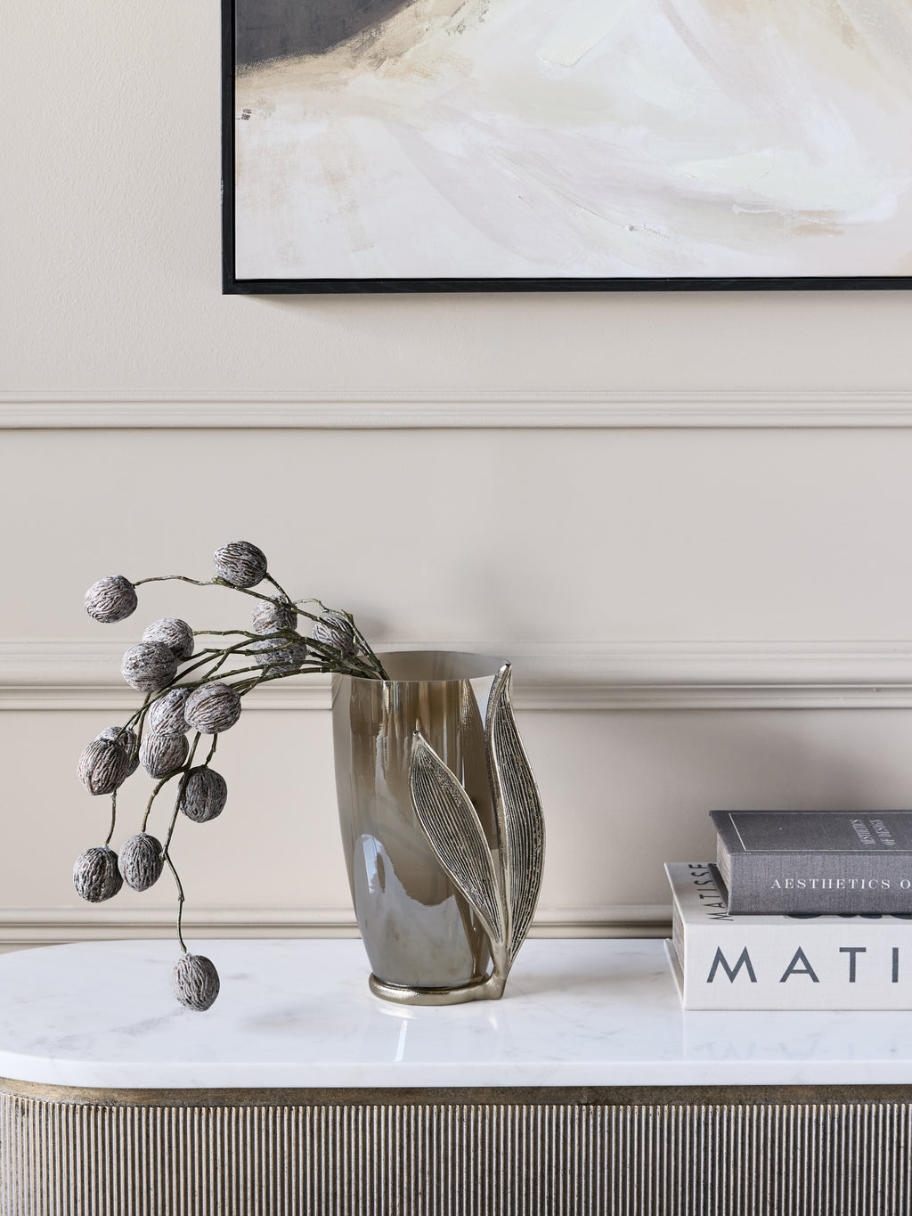 Leaf vase holding decorative pod stems on a pale console beside stacked books, beneath abstract artwork.
