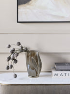 Leaf vase holding decorative pod stems on a pale console beside stacked books, beneath abstract artwork.
