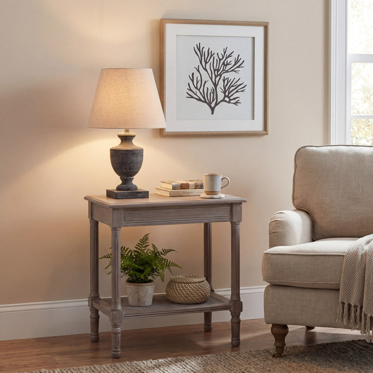 Urn Plinth Table Lamp shown lit on a side table beside books and a mug, with a cream armchair and framed branching print nearby.