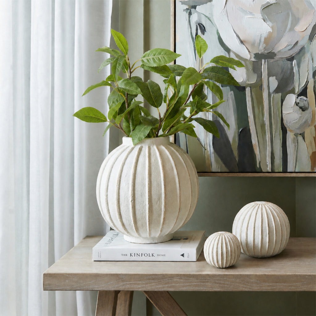 Laurel-style foliage arranged in a round ribbed white vase on a book, beside two ribbed ornaments on a wooden console with floral artwork behind.