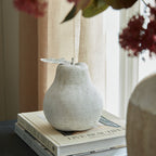 Pale pear ornament on stacked books beside a window with beige curtains, with its leaf extending to the left.