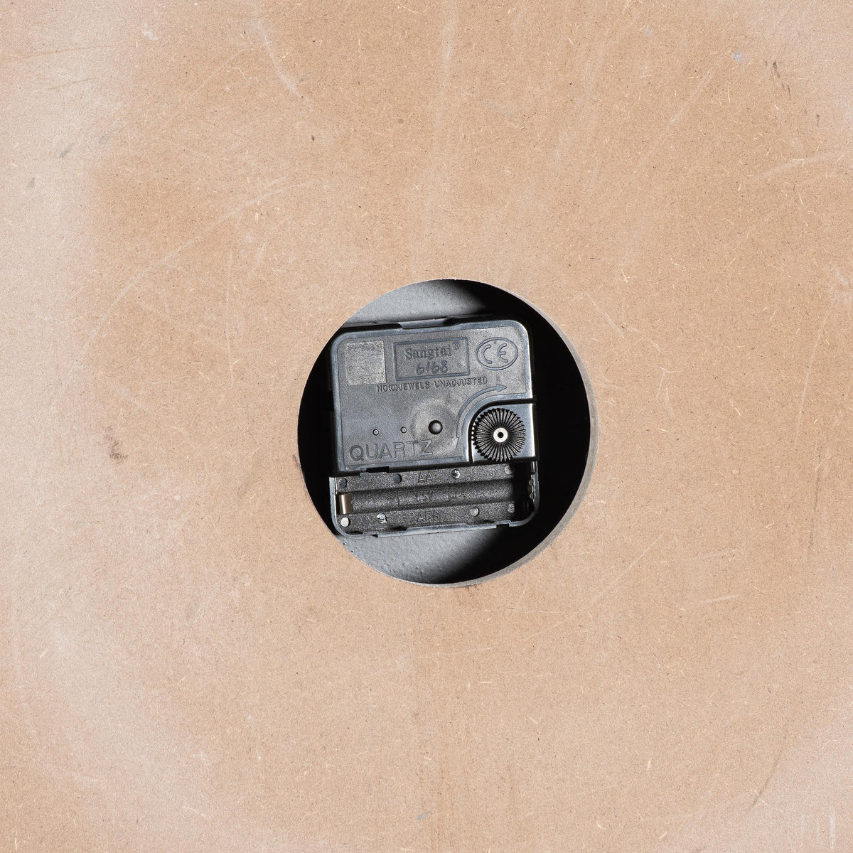 Rear clock detail showing a black movement, adjustment wheel and empty battery compartment through a round cutout in the brown backing.