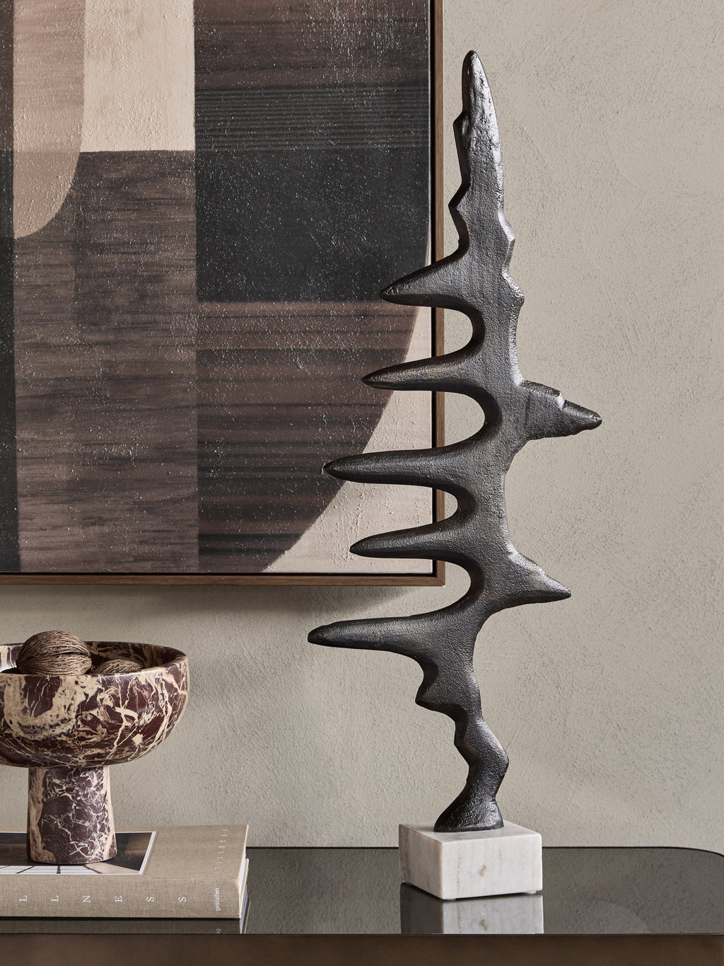 Waveform Sculpture on a reflective console beside books and a patterned pedestal bowl, with abstract artwork behind.