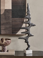 Waveform Sculpture on a reflective console beside books and a patterned pedestal bowl, with abstract artwork behind.
