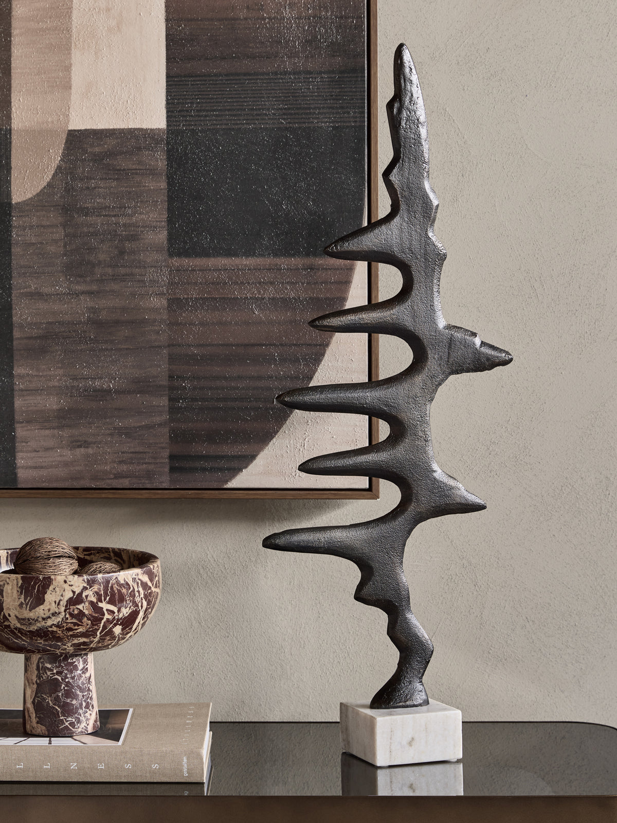 Waveform Sculpture on a reflective console beside books and a patterned pedestal bowl, with abstract artwork behind.